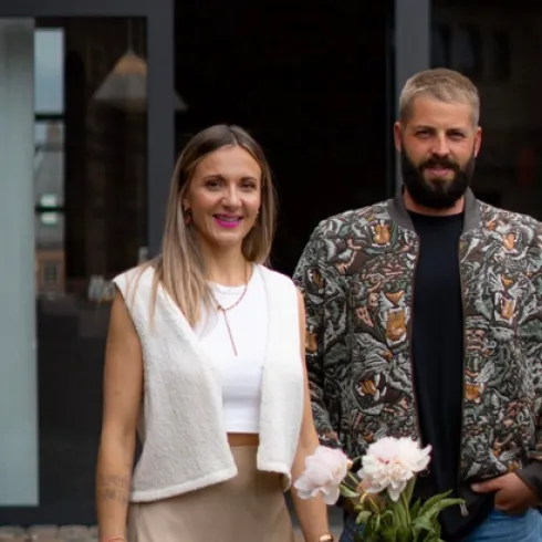 Ein Paar steht lächelnd vor einem modernen Hintergrund. Sie tragen modische Kleidung und es sind einige Pflegeprodukte und Blumen auf einem Tisch platziert.