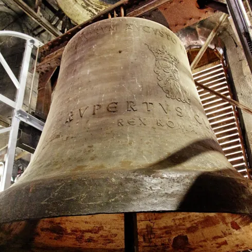 Eine große, alte Glocke aus Metall steht im Vordergrund. Im Hintergrund sind Teile des Glockenturms sichtbar.