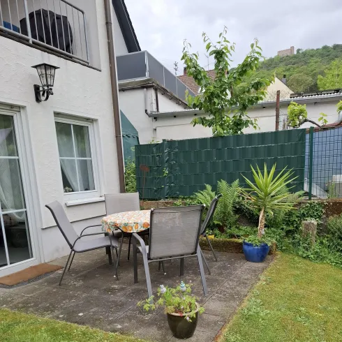 Ein gemütlicher Gartenbereich mit einem Tisch und Stühlen auf einer Terrasse. Umgeben von Pflanzen und einer grünen Wiese.
