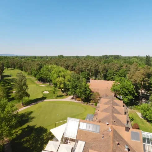 Eine weitläufige Grünlandschaft mit Bäumen und einem Golfplatz. Der Himmel ist klar und blau, was eine angenehme Atmosphäre schafft.
