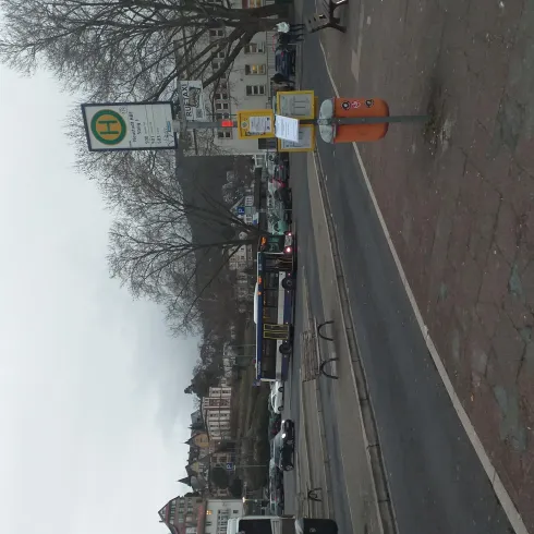 Eine Haltestelle an einer Straße mit einem Warteschild und wenigen Fahrzeugen. Im Hintergrund sind Gebäude und Bäume zu sehen.