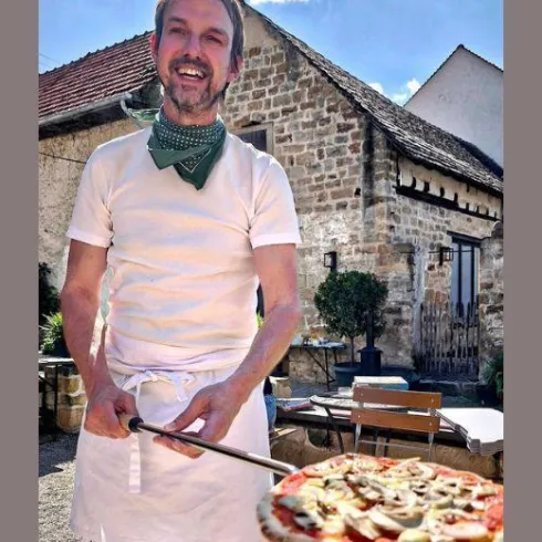 Ein jovialer Mann in Kochkleidung steht draußen und hält eine Pizzaschaufel mit einer frisch zubereiteten Pizza. Im Hintergrund ist ein charmantes Gebäude und ein gemütlicher Außenbereich zu sehen.