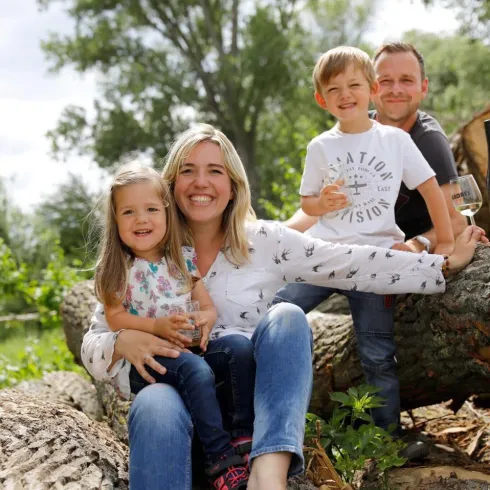 Eine glückliche Familie sitzt auf Baumstämmen im Freien. Die Kinder halten Gläser in den Händen und lächeln in die Kamera.