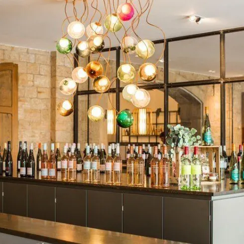 Ein moderner Weinsalon mit einer Vielzahl von Weinflaschen auf einer dunklen Theke. Über der Theke hängen stilvolle, bunte Lampen.