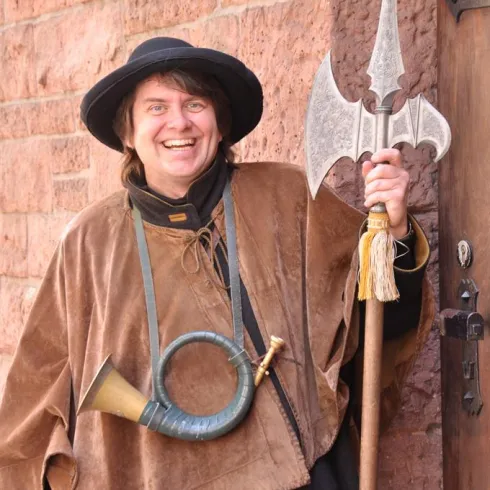 Ein Mann in historischem Gewand steht vor einer Wand und lächelt. Er hält einen Speer und ein Horn in der Hand.