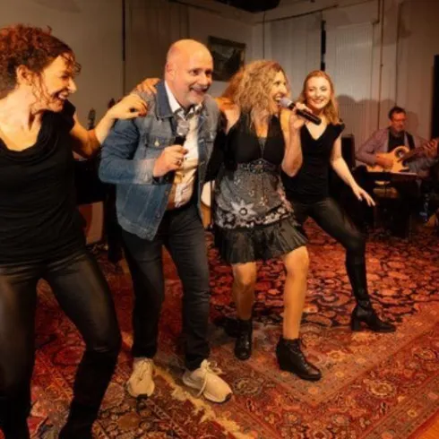 Eine fröhliche Bühnenaufführung mit vier Personen, die singen und tanzen. Im Hintergrund spielt eine Band Musik.
