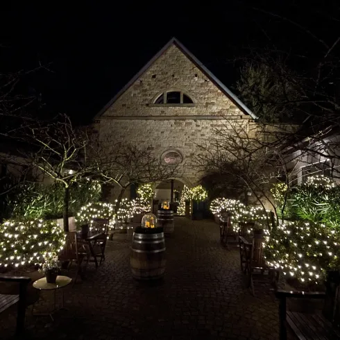 Eine stimmungsvolle, beleuchtete Gartenanlage bei Nacht mit Bäumen und Sitzgelegenheiten. Das Gebäude im Hintergrund hat eine charmante Steinfassade.