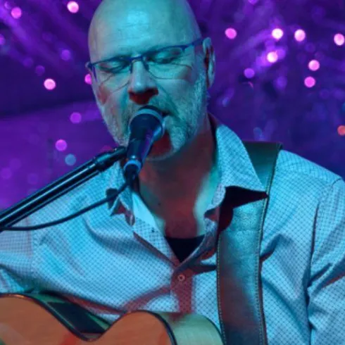 Ein Musiker spielt Gitarre und singt in einer bunten Umgebung. Im Hintergrund sind funkelnede Lichter zu sehen.