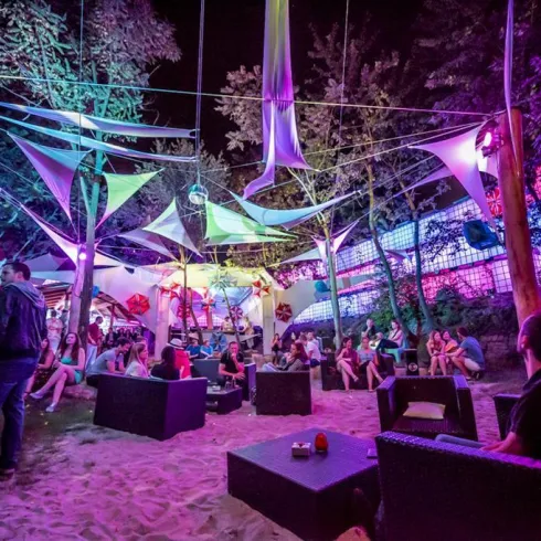 Eine lebhafte Nachtveranstaltung im Freien mit bunten Lichtern und vielen Gästen. Die entspannte Atmosphäre wird durch überdachte Sitzbereiche und dekorative Elemente verstärkt.