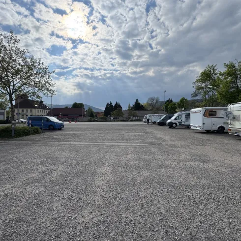 Ein großer, leerer Parkplatz mit mehreren Campingfahrzeugen und Bäumen. Der Himmel ist bewölkt, und die Umgebung wirkt ruhig.