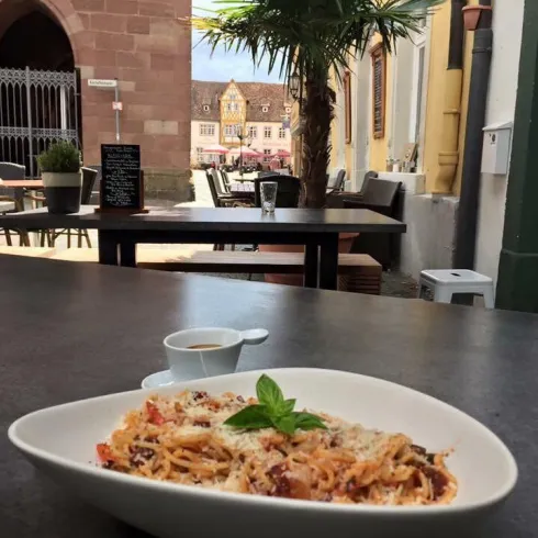 Ein Teller Pasta mit Tomaten und Käse steht auf einem Tisch. Im Hintergrund sieht man eine gemütliche Umgebung mit Pflanzen und Tischen.