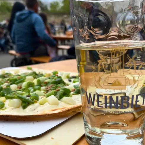 Ein Glas Wasser steht neben einer Pizza mit frischem Gemüse auf einem Holztisch. Im Hintergrund sind Menschen zu sehen, die sich im Freien entspannen.