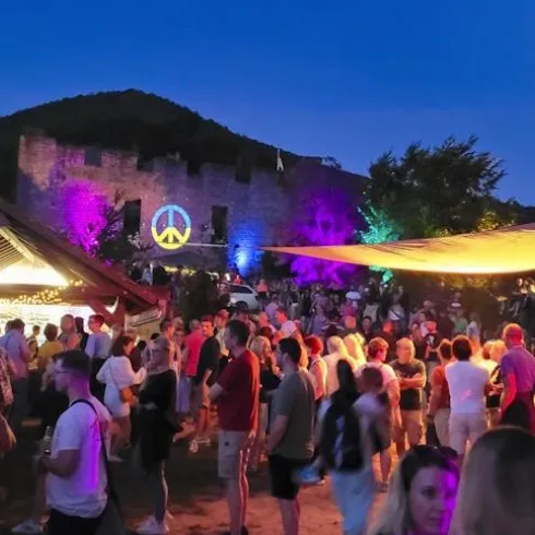 Eine lebhafte Veranstaltung bei Nacht mit vielen Menschen und bunten Lichtern. Im Hintergrund ist eine historische Burg mit einem Friedenszeichen beleuchtet zu sehen.