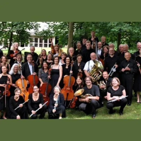 Eine große Gruppe von Orchestermusikern steht im Freien unter Bäumen. Die meisten tragen schwarze Kleidung und lächeln in die Kamera.