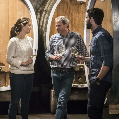 Drei Personen stehen in einem Weinkeller und halten Gläser. Im Hintergrund sind große Holzfässer zu sehen.