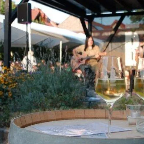 Eine gemütliche Außenveranstaltung mit Live-Musik. Im Vordergrund stehen zwei Gläser Wein auf einem Fass, umgeben von blühenden Blumen.