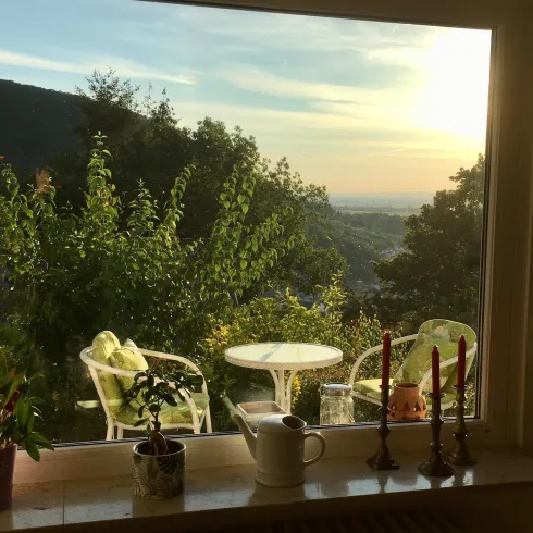 Ein schöner Ausblick auf eine grüne Landschaft mit einem Tisch und Stühlen im Freien. Im Vordergrund stehen einige Pflanzen und Kerzen auf der Fensterbank.