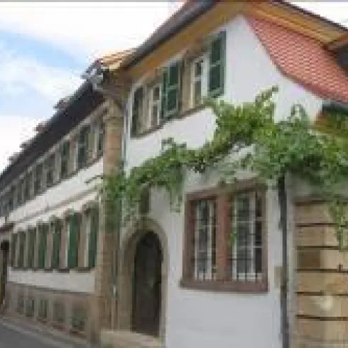 Ein charmantes, historisches Gebäude mit weißen Wänden und grünen Fensterläden. Pflanzen ranken an der Fassade und verleihen dem Haus einen einladenden Charakter.