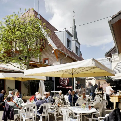 Ein gemütliches Café im Freien mit vielen Tischen und Stühlen. Menschen genießen die Sonne und die entspannte Atmosphäre.