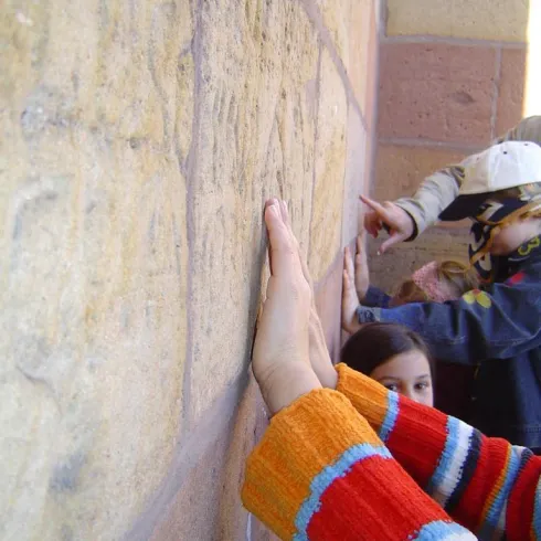 Eine Gruppe von Kindern berührt eine Wand aus Stein. Ein Mädchen mit einem bunten Pullover ist im Vordergrund zu sehen.