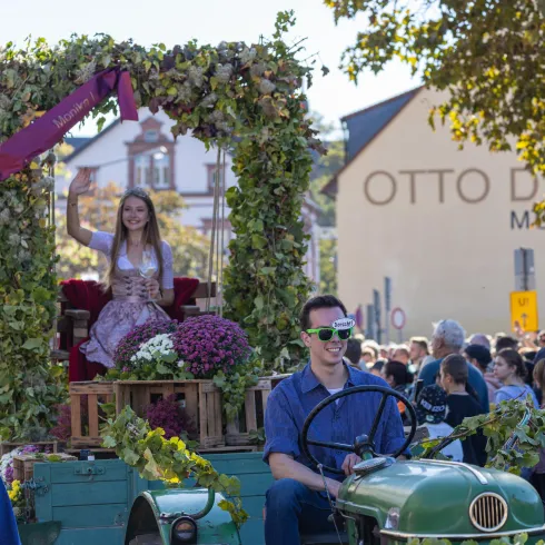 Eine festliche Parade mit einem geschmückten Traktor. Eine junge Frau sitzt auf dem Wagen und wird von einer Menschenmenge umgeben.