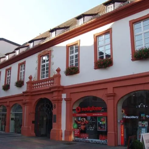 Ein historisches Gebäude mit rotem Putz und großen Fenstern. Im Erdgeschoss sind Geschäfte wie Vodafone und Blackberry zu sehen.