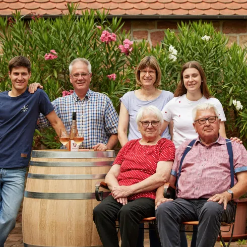 Eine Familiengruppe steht vor einem Weinfass und blühenden Pflanzen. Alle lächeln und wirken freundlich.