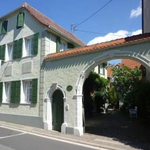 Ein charmantes Haus mit grünen Fensterläden und einem roten Dach. Der Eingang wird von einem geschwungenen Torbogen flankiert, der in einen grünen Innenhof führt.