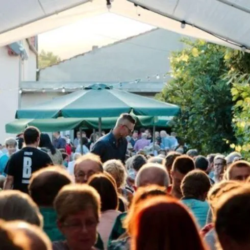 Eine belebte Veranstaltung mit vielen Menschen unter einem Zelt. Im Hintergrund sind Bäume und Sonnenschein zu sehen.