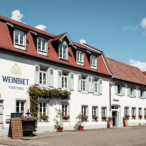 Ein einladendes Weingut mit einer hellen Fassade und einem roten Dach. Blumen schmücken die Fenster, und der Himmel ist blau mit wenigen Wolken.
