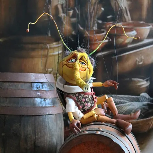 Eine Marionette mit einem fröhlichen Gesicht sitzt auf einem Holzfass. Im Hintergrund sind traditionelle Holzgegenstände zu sehen.