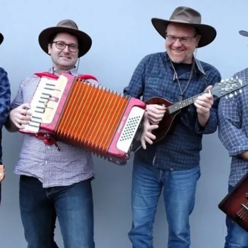Vier Musiker tragen Hüte und stehen lächelnd zusammen. Einer von ihnen spielt ein Akkordeon, während die anderen verschiedene Instrumente halten.