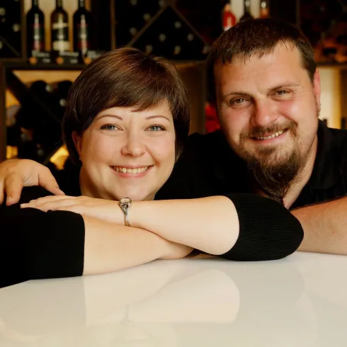 Ein Paar lächelt freundlich in einem Weinkeller. Im Vordergrund steht eine Flasche Wein.