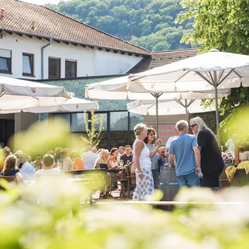 Eine belebte Terrasse mit vielen Gästen unter Sonnenschirmen. Im Hintergrund sind Gebäude und die grüne Landschaft sichtbar.