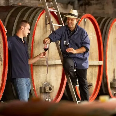 Zwei Männer probieren Wein in einem Weinkeller. Sie stehen neben großen Holzfässern und halten Weingläser in der Hand.