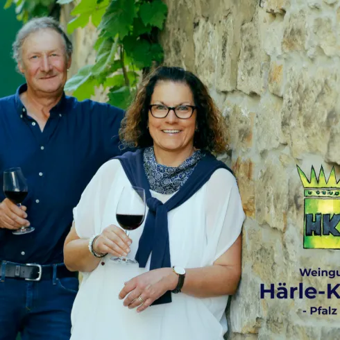 Ein Paar posiert vor einer Steinewand, jeder hält ein Glas Wein. Der Hintergrund zeigt Weinreben und den Namen des Weinguts "Härle-Kerth" aus der Pfalz.