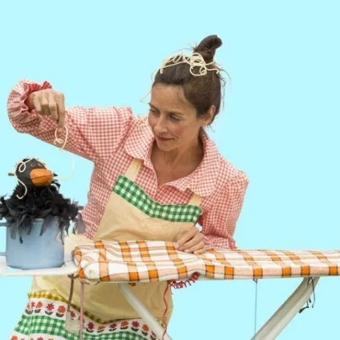 Eine Frau spielt mit einer Puppenfigur, die einen Vogel darstellt. Sie sitzt an einem Tisch, auf dem ein Bügeltuch liegt.