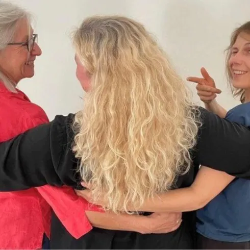 Drei Frauen stehen auf einer Bühne und halten Skripte in der Hand. Im Hintergrund sind Tische, Stühle und ein Musikinstrument zu sehen.