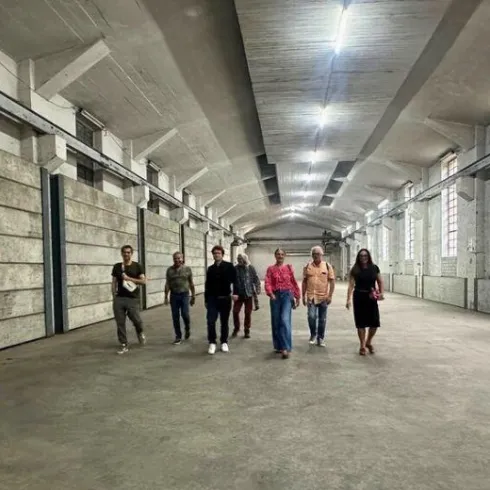 Eine Gruppe von Menschen geht durch einen großen, leeren Raum mit hohen Decken und Betonwänden. Das Licht kommt von den Fenstern und den Deckenlampen.