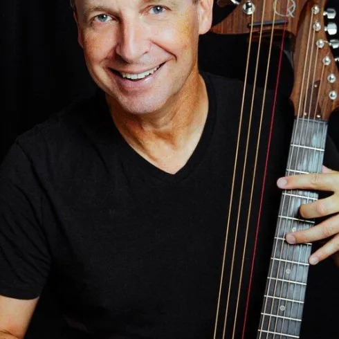 Ein lächelnder Mann hält eine Gitarre in der Hand. Er trägt ein schwarzes T-Shirt vor einem dunklen Hintergrund.