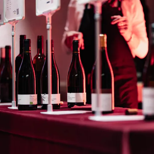 Eine lange Theke mit verschiedenen Flaschen Wein steht im Vordergrund. Im Hintergrund ist eine Person mit einem weißen Hemd zu sehen, die sich um die Weine kümmert.