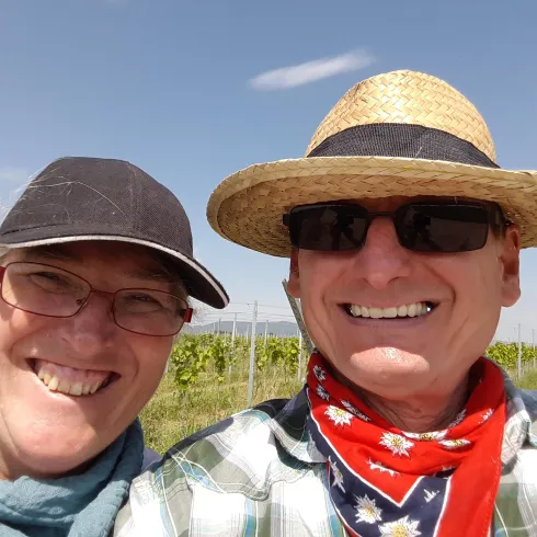 Ein Paar lächelt in einem Weinberg. Sie tragen Sonnenbrillen und einen Strohhut, während der Himmel klar ist.