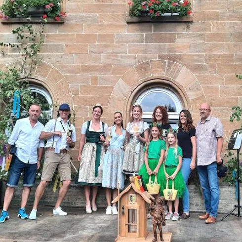 Eine Gruppe von Menschen in traditioneller Tracht steht vor einer dekorierten Wand. Es gibt mehrere Frauen und Kinder, die fröhlich posieren.