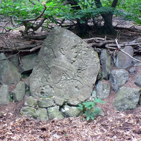 Ein kunstvoll behauener Stein mit einer Büste inmitten eines Waldes. Der Stein ist von einem Steinhügel und grünen Pflanzen umgeben.