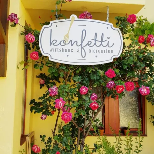 Ein Schild mit der Aufschrift "konfetti" hängt an einer blühenden Rosenhecke. Im Hintergrund sieht man ein Fenster eines Gebäudes.