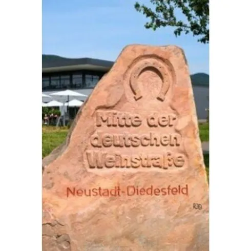 Ein Wegweiser aus Stein mit der Aufschrift "Mitte der deutschen Weinstraße". Im Hintergrund sind Bäume und ein Gebäude sichtbar.