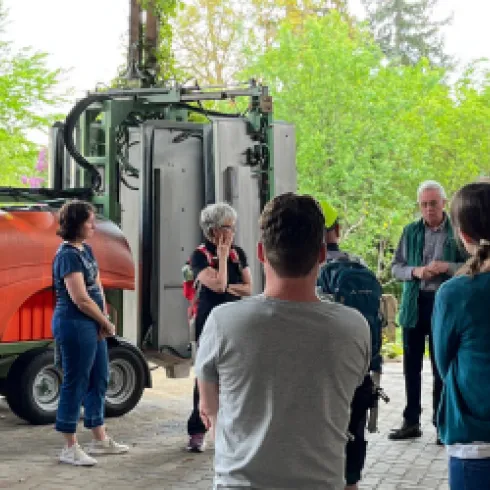 Eine Gruppe von Menschen hört einem Vortrag in einem Garten oder Hof zu. Im Hintergrund steht ein Traktor mit einer großen orangefarbenen Anlage.