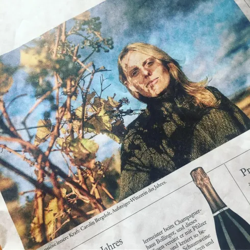 Eine Winzerin steht zwischen Weinreben und lächelt in die Kamera. Die Szene vermittelt eine warme, herbstliche Atmosphäre.