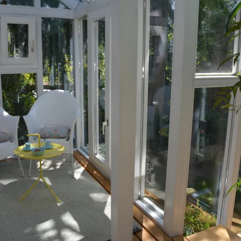 Eine helle Veranda mit zwei weißen Stühlen und einem kleinen gelben Tisch. Viele Pflanzen und Licht durch die Fenster schaffen eine entspannte Atmosphäre.