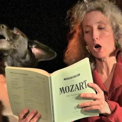 Eine Frau singt laut und hält ein Notenbuch mit dem Titel "Mozart". Neben ihr steht ein Hund, der ebenfalls den Kopf in die Höhe richtet.